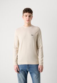 Beige pitkähihaine villapaita pyöreällä kauluksella, jossa on hienovarainen röyhelöpinnanne ja pieni musta "BOSS" -logo rinnassa. Yhdistetty sinisiin farkkuihin.