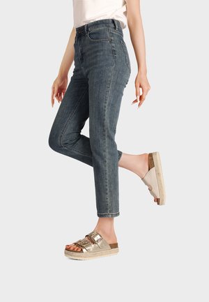Donna con jeans blu a vita alta e sandali beige con fibbia, in piedi con una gamba sollevata su uno sfondo semplice.