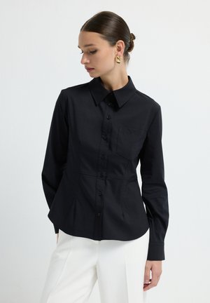 Woman wearing black long-sleeve button-up shirt with collar and chest pocket, paired with white pants, looking to her left with hair in a bun.