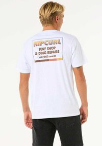 T-shirt bianca a maniche corte con una grafica colorata sul retro che presenta "RIPCURL" e testo per un negozio di surf, stampato con diversi font.
