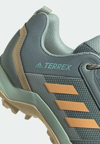 Chaussure de randonnée bleu clair avec dessus en maille, semelle en caoutchouc et accents bruns. Présente trois bandes orange et un logo vert sur le côté.