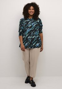 Camicia nera con motivo astratto blu e verde, design a bottoni, maniche a tre quarti; abbinata a pantaloni beige a sigaretta e scarpe nere.