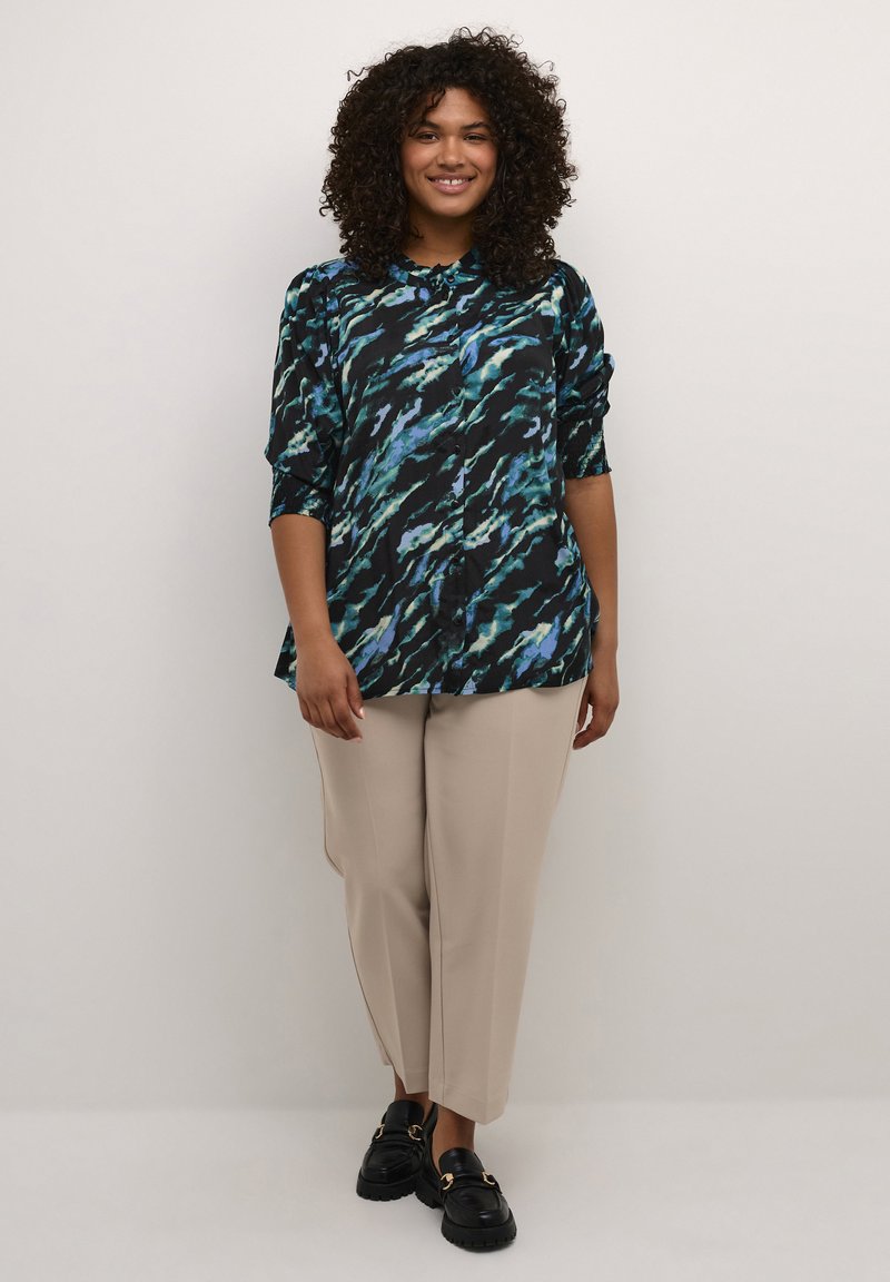 Camicia nera con motivo astratto blu e verde, design a bottoni, maniche a tre quarti; abbinata a pantaloni beige a sigaretta e scarpe nere.
