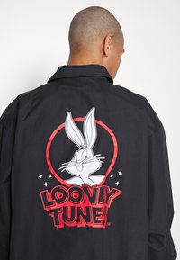 Person med kort hår som bär en svart jacka med ett Bugs Bunny-tecknat motiv och en röd "Looney Tunes"-logotyp på ryggen mot vit bakgrund.