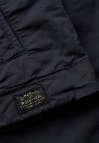 Marineblåt stof med en glat tekstur har en fremtrædende sort label med teksten "Superdry", placeret på en syet kant.