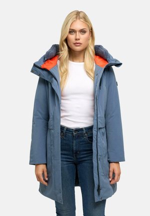 Lyshåret kvinde iført en blå lang vinterfrakke med orange og marineblå imiteret pelsfor, hvid skjorte og blå jeans, vender fremad med neutralt ansigtsudtryk.