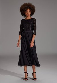 Robe midi noire à manches trois-quarts ; corsage à sequins, jupe fluide et lisse, taille marquée, associée à des talons noirs �à lanières.