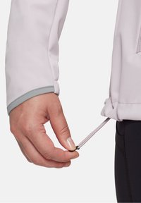 Ljuslila softshell-jacka med gråa detaljer, som visar en hand som justerar den elastiska snöret i nederkanten för en säker passform.