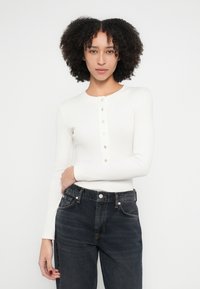 Balts garroku tops ar apaļu kakla izgriezumu un piecu pogu rāvējslēdzēju, kombinēts ar tumši ziliem džinsiem ar augstu vidukli. Gluds audums, pieguļošs dizains.