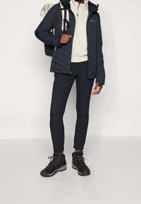 Giacca nera isolata con design trapuntato, pullover crema, pantaloni scuri e scarponi da trekking neri. Il modello porta uno zaino beige e nero.