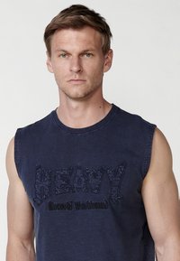 Mornarsko modra brezrokavnik iz mehkega blaga, ki ima veliko vezeno besedilo "HEAVY" in pod njim napis "KoroShi Washbrand" v črni barvi.