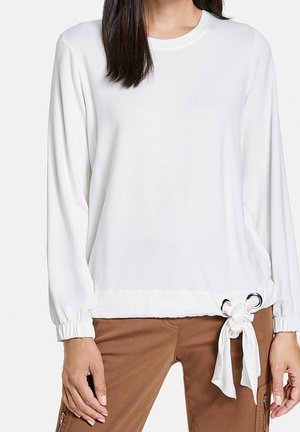Femme portant un haut blanc à manches longues avec des poignets élastiques et un détail de nœud noué avec des œillets métalliques à l'ourlet, associé à un pantalon marron.