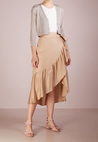 Ljust grå, glittrande cardigan över en vit topp, i kombination med en beige, knälång wrapkjol med en volangslå. Ljust rosa sandaler med remmar.