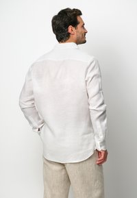 Camisa de lino blanca con un cuello clásico, mangas largas y un dobladillo curvado. La parte trasera presenta una textura suave y ligera.