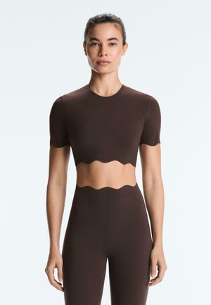 Femeie care poartă un crop top maro închis cu mâneci scurte și colanți cu talie înaltă și margini ondulate.