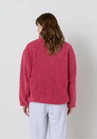 Roza jakna iz puhastih flis materialov z visokim ovratnikom, širokim krojem in rebrastimi manšetami, kombinirana z sivimi športnimi hlačami. Prikaz od zadaj.