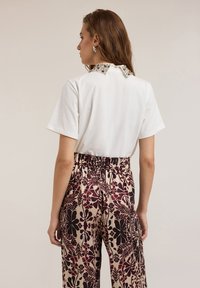 Camisa blanca de manga corta con cuello adornado, combinada con pantalones holgados de cintura alta que presentan un gran estampado floral en burdeos y crema.