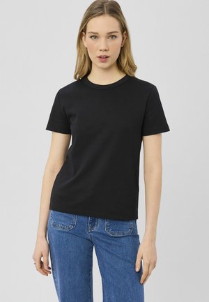 Jeune femme aux longs cheveux blonds portant un t-shirt noir à manches courtes et un jean bleu, debout devant un fond uni gris clair.