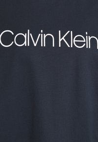 Námorně modré bavlněné tričko s velkým bílým logem "Calvin Klein" v moderním bezserifovém písmu přes hruď. Hladká textura.
