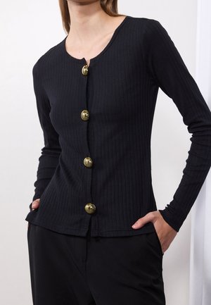 Femme portant un haut noir côtelé à manches longues avec de gros boutons ronds dorés, les mains dans les poches d’un pantalon noir.