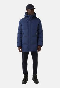 Mørkeblå pufferjakke med hætte, lynlåslommer og quiltet design. Kombineret med mørke bukser og sorte høj-top sneakers.