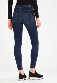 Mörkblå skinny jeans med hög midja, med två bakfickor och rå fåll. Bärs med svarta skor utan snörning.