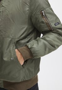 Olive green bomber jacket with a smooth, shiny texture; features ribbed cuffs, a zippered pocket on the arm, and side pockets.
