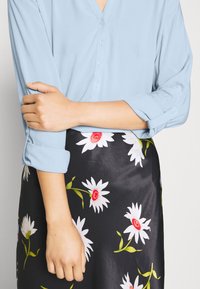 Blouse bleu clair à boutons avec un décolleté en V et des manches retroussées, associée à une jupe noire ornée de marguerites blanches et de centres rouges.