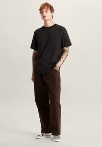 Black short-sleeve t-shirt, brown corduroy pants, and black-and-white checkered slip-on shoes; tattoos visible on arms.