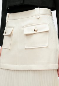 Gonna in tessuto crema con due tasche esterne dotate di bottoni dorati e sezione inferiore plissettata, progettata con una cinta in vita sagomata.