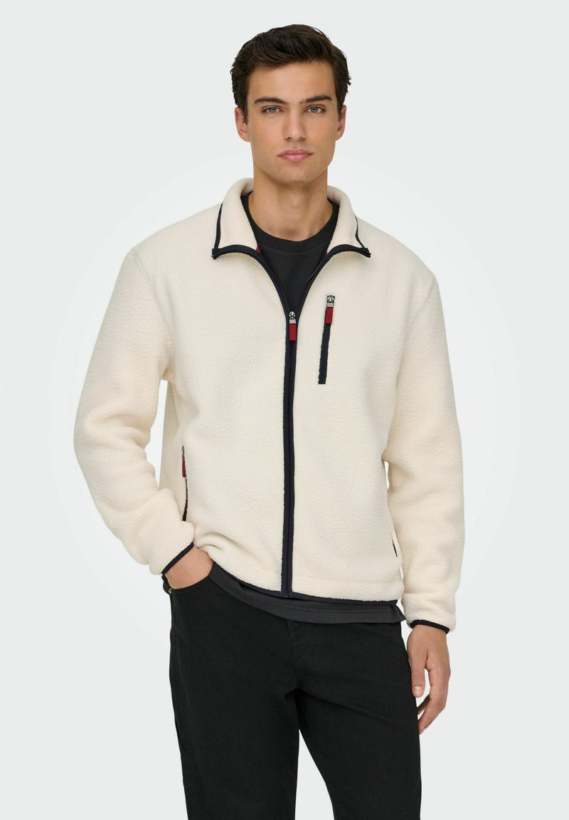 Mann in einer hellbeigen Fleecejacke mit Reißverschluss und schwarzen Akzenten sowie roten Zip-Ziehern, schwarzem Hemd darunter und schwarzen Hosen, steht mit einer Hand in der Tasche.