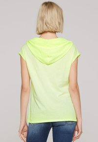 Neon yellow short-sleeve hoodie with a drawstring, featuring a relaxed fit and soft texture, paired with blue jeans.