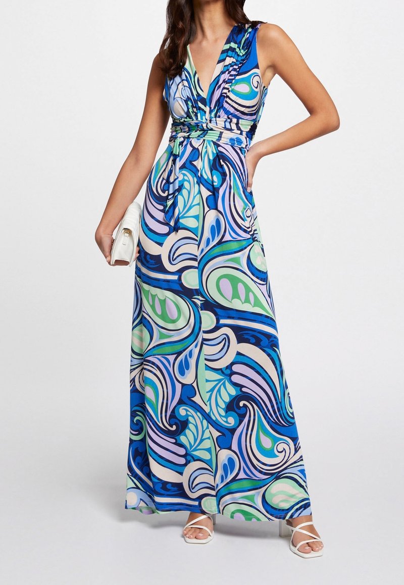 Žena v bezramenných, výstřihových šatech až na podlahu s modrými, fialovými, zelenými a bílými vířícími vzory, držící bílou kabelku a obutá v bílých sandálech.