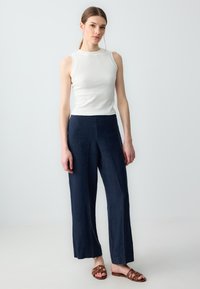 Witte geribde mouwloze top, gecombineerd met donkerblauwe wijde broek met een gladde textuur en een vlakke voorkant. Bruine sandalen met banden.