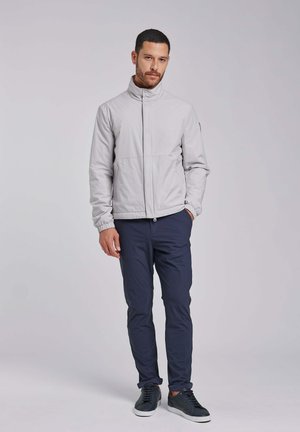 Uomo con giacca grigio chiaro con cerniera, pantaloni blu navy e scarpe da ginnastica nere, in piedi con una mano in tasca su sfondo semplice.