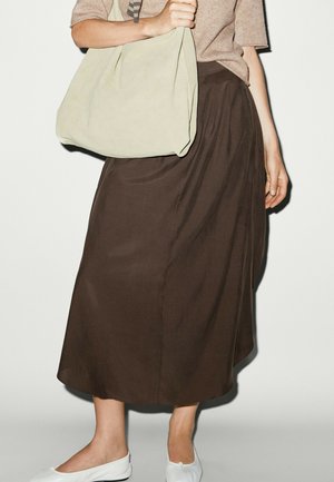 Person trägt einen braunen Midirock, beiges Oberteil, weiße Ballerinas und trägt eine große hellbeige Tote-Tasche über einer Schulter.