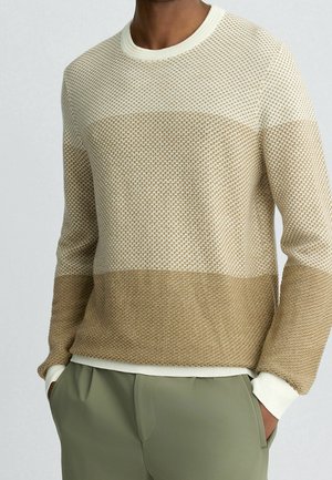 Uomo che indossa un maglione beige e crema a maglia strutturata con polsini e orlo a coste, abbinato a pantaloni verde oliva, mani nelle tasche.