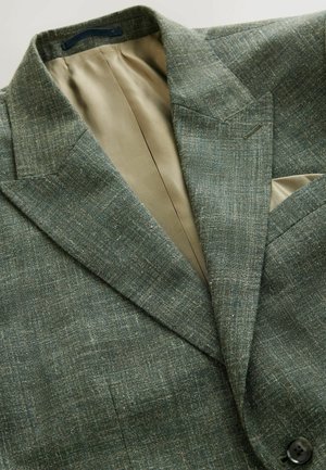 Zielony tweedowy blazer z subtelnie wplecionym wzorem w kratę, z klapami w kształcie notcha i jedwabistą beżową podszewką. Posiada poszetkę oraz czarny guzik.