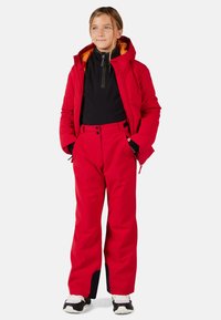Giacca isolante rossa con cappuccio, abbinata a pantaloni da sci rossi e una maglietta nera. Caratterizzata da cerniere, tasche e accenti a contrasto.