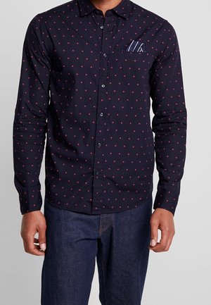 Homme portant une chemise bleu marine à boutons avec un motif en losanges rouges et un mouchoir de poche rayé, assortie à un jean bleu foncé.