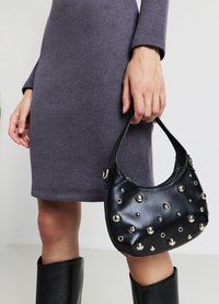 Borsa a mano in pelle nera con borchie e chiodi in argento, forma curva e manico corto, tenuta in una mano accanto a un vestito grigio e stivali neri.
