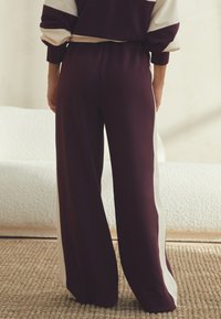 Široke hlače bordo barve z kremnimi stranskimi črtami, iz mehkega materiala. Imajo elastičen pas in sproščen kroj, zasnovane za udobje.