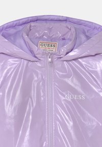Chaqueta con capucha de color púrpura claro y brillante, con cremallera central, logo "GUESS" en el pecho y etiqueta de la marca en el interior del cuello.