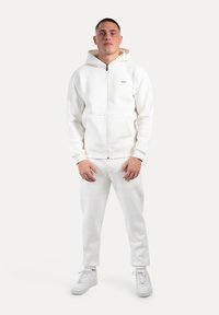 Felpa bianca con zip e cappuccio, con un piccolo logo sul petto. Abbinata a pantaloni della tuta bianchi e sneakers bianche.