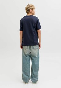 Έφηβος με κοντά ξανθά μαλλιά, φορώντας μπλε σκούρο T-shirt και ανοιχτό μπλε τζιν με φαρδιά πόδια, γυρισμένος προς τα πίσω σε απλό φόντο.