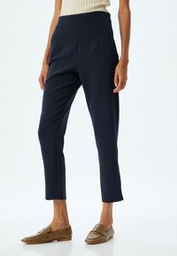 Navyblauwe broek met plooien aan de voorkant, taps toelopende pijpen en subtiele zijsplitten aan de zoom. Gemaakt met bruine, gemarmerde instapschoenen.