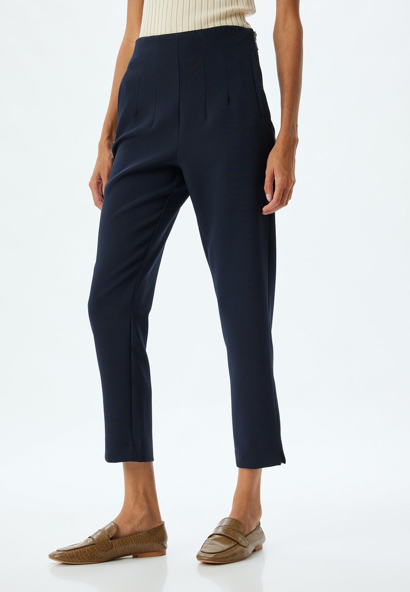 Navyblauwe broek met plooien aan de voorkant, taps toelopende pijpen en subtiele zijsplitten aan de zoom. Gemaakt met bruine, gemarmerde instapschoenen.