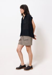 Schwarze, locker sitzende Bluse mit kurzen Ärmeln aus strukturiertem Stoff; kombiniert mit beige-schwarz karierten Shorts und schwarzen Schnürschuhen.