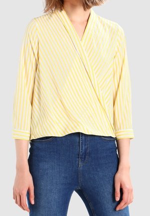 Woman wearing yellow and white diagonal striped wrap blouse with three-quarter sleeves and blue jeans, standing against plain background.