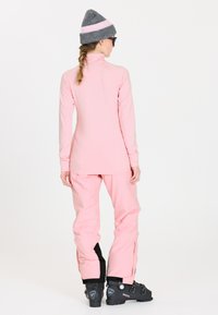 Rosa Langarmshirt und passende rosa Hose, aus glattem Stoff. Eine graue gestreifte Mütze und schwarze Schneestiefel vervollständigen das Outfit.
